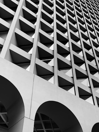 A striking architectural facade characterized by a geometric pattern of recessed square openings. The structure is composed of grid-like concrete beams, creating a sense of depth and rhythm. Arched sections can be seen at the bottom, contributing to the overall modernist aesthetic.