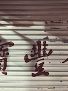 A metal roller shutter with peeling paint and faded red characters painted across it. The surface shows signs of wear and aging, giving a rustic appearance.