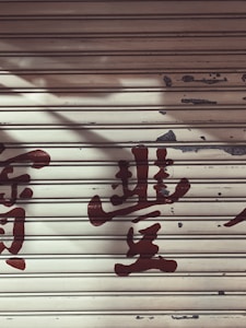 A metal roller shutter with peeling paint and faded red characters painted across it. The surface shows signs of wear and aging, giving a rustic appearance.
