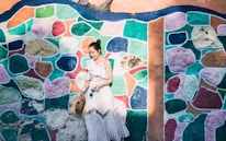 A woman in a white, polka-dotted dress with off-shoulder design is standing in front of a brightly colored mosaic wall. She is holding a woven handbag and looks down with a gentle smile. The wall features a patchwork of vibrant colors including green, pink, blue, and orange, creating a lively and artistic background.