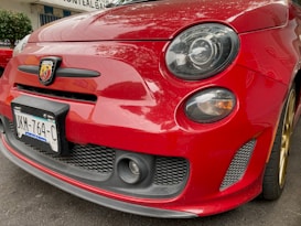 A close-up view of the front end of a red Fiat Abarth car, showcasing the distinctive Abarth emblem on the grille. The vehicle's round headlight and fog light are clearly visible, along with a portion of the hood and the front license plate bearing a number 'UKM-764-C'. The car is parked on a paved street next to greenery.