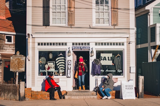 A small boutique with a sign reading 'Wicked Peacock' has a window display featuring jewelry and clothing items. Two people sit outside the shop, and another person stands in the doorway. The shop facade is painted white with large glass windows advertising a sale.