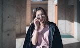 A young woman with light brown hair is wearing sunglasses and a light pink dress under a black jacket. She stands in a sunlit concrete hallway, holding a smartphone to her ear, casting soft shadows on the walls around her.