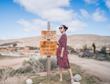 A woman in a red polka dot dress stands in a desert landscape next to a wooden sign with a humorous message about driving. The background features dry plants, a dirt road, and distant hills under a blue sky with scattered clouds.