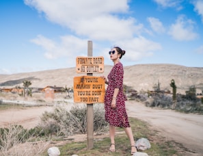 A woman in a red polka dot dress stands in a desert landscape next to a wooden sign with a humorous message about driving. The background features dry plants, a dirt road, and distant hills under a blue sky with scattered clouds.