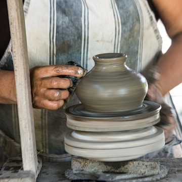 A person is meticulously shaping a clay pot on a potter's wheel. Their hands, coated in clay, gently mold the spinning vessel. The wheel and the pot are in motion, suggesting the dynamic process of pottery.