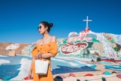 A woman wearing a bright yellow dress and sunglasses stands with a white bag in front of a colorful, painted hillside with the words 'God is Love' and a large cross on top. The landscape features vivid pastel colors and a clear blue sky, creating a vibrant and artistic display.