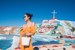A woman wearing a bright yellow dress and sunglasses stands with a white bag in front of a colorful, painted hillside with the words 'God is Love' and a large cross on top. The landscape features vivid pastel colors and a clear blue sky, creating a vibrant and artistic display.