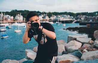 A person is taking a photo with a camera while standing on rocky terrain by the waterfront. Several boats are visible floating in the water, and a quaint town with various buildings is seen in the background. The scene captures a mix of natural and urban elements.