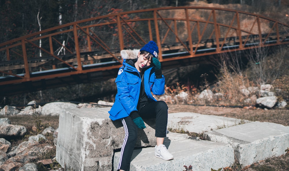 man in blue jacket and black pants sitting on concrete wall during daytime