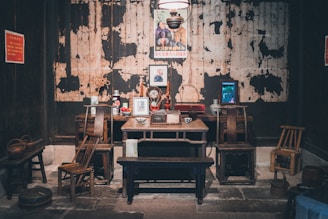 A vintage room with a rustic setup featuring a wooden table and chairs. There's an assortment of objects on the table including framed pictures, a ceramic mug, and decorative items. The backdrop reveals a worn, textured wall with a poster featuring two figures. Additional items such as small stools and a woven basket are scattered around.