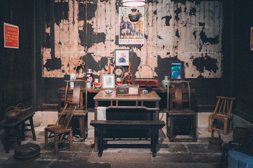 A vintage room with a rustic setup featuring a wooden table and chairs. There's an assortment of objects on the table including framed pictures, a ceramic mug, and decorative items. The backdrop reveals a worn, textured wall with a poster featuring two figures. Additional items such as small stools and a woven basket are scattered around.