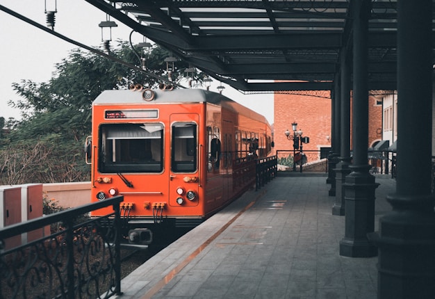 A vintage-style orange train is stationed on a platform with ornate railings and overhead structures. The setting includes a lamp post and a background of trees and brick buildings, creating a nostalgic atmosphere.