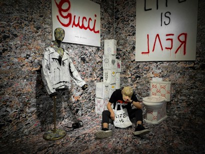 A room with walls and floor covered in a collage-like pattern hosts a mannequin wearing a distressed white jacket. Next to it, a person wearing a hat with a face design sits on the floor, leaning against a sack labeled 'LIFE IS'. The setup includes several cylindrical objects wrapped in materials with various patterns, and a toilet emblazoned with red 'GG' logos. On the wall, a large sign reads 'Gucci' and another sign features the text 'LIFE IS ∃AL'.
