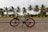 A bicycle is parked on a roadside with palm trees and a large white building in the background. The setting appears to be tropical with cloudy skies and a scenic landscape.