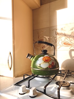 A green kettle with floral and butterfly designs sits on a white gas stove. The kettle has a black handle and knob. Sunlight is streaming in, casting shadows and illuminating the wall's decorative tiles with floral patterns in the background. The stove has two visible white knobs in the foreground.