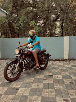 A person wearing a colorful helmet and blue shirt is sitting on a black Royal Enfield motorcycle. The scene is set in a paved area with a backdrop of lush green trees and a blue wall.