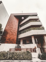 A modern multi-level building featuring a combination of wood and concrete materials. The facade includes large, rectangular balconies adorned with lush greenery, providing a natural contrast to the geometric architecture. A well-maintained garden area with shrubs is visible at the front, along with a wooden fence and a sidewalk leading to the entrance.