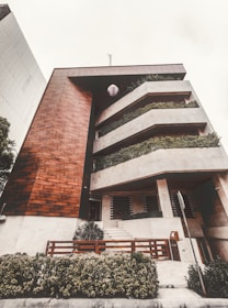 A modern multi-level building featuring a combination of wood and concrete materials. The facade includes large, rectangular balconies adorned with lush greenery, providing a natural contrast to the geometric architecture. A well-maintained garden area with shrubs is visible at the front, along with a wooden fence and a sidewalk leading to the entrance.