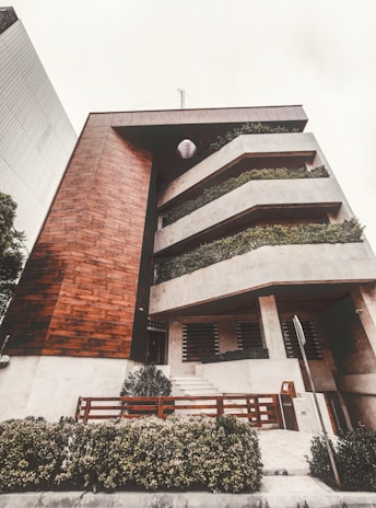 A modern multi-level building featuring a combination of wood and concrete materials. The facade includes large, rectangular balconies adorned with lush greenery, providing a natural contrast to the geometric architecture. A well-maintained garden area with shrubs is visible at the front, along with a wooden fence and a sidewalk leading to the entrance.