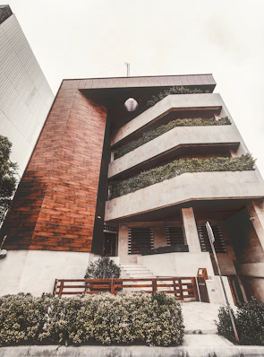A modern multi-level building featuring a combination of wood and concrete materials. The facade includes large, rectangular balconies adorned with lush greenery, providing a natural contrast to the geometric architecture. A well-maintained garden area with shrubs is visible at the front, along with a wooden fence and a sidewalk leading to the entrance.