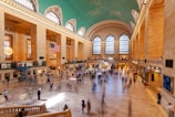 A bustling indoor train terminal with high arched windows and a distinctive turquoise ceiling decorated with constellations. An American flag is prominently displayed in the middle, hanging from the ceiling. The terminal is full of people walking around and standing in various areas. There are ticket counters and information screens along the left side. The floor is made of polished stone tiles, and the space is brightly lit by both natural and artificial lighting.