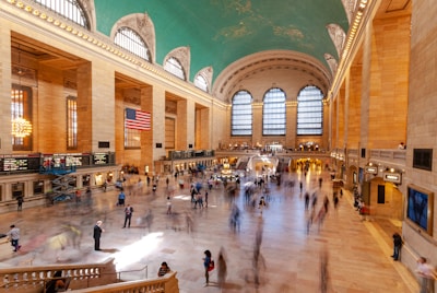A bustling indoor train terminal with high arched windows and a distinctive turquoise ceiling decorated with constellations. An American flag is prominently displayed in the middle, hanging from the ceiling. The terminal is full of people walking around and standing in various areas. There are ticket counters and information screens along the left side. The floor is made of polished stone tiles, and the space is brightly lit by both natural and artificial lighting.