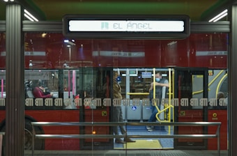 A city bus with a sign indicating the destination 'El &Aacute;ngel' is seen through a glass panel. Inside, a couple of seated passengers are visible, with one resting and another standing near the door. The scene is illuminated by artificial lighting, reflecting an urban ambiance.