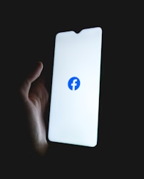 A hand holding a smartphone with a brightly lit screen displaying the Facebook logo in the center.