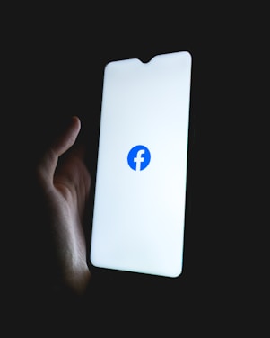 A hand holding a smartphone with a brightly lit screen displaying the Facebook logo in the center.
