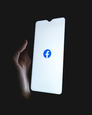 A hand holding a smartphone with a brightly lit screen displaying the Facebook logo in the center.
