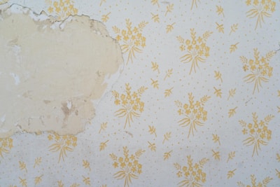 A section of wall features peeling wallpaper with a faded floral pattern. The wallpaper has a beige background with yellow floral designs. Part of the wall is exposed, revealing an uneven, discolored surface underneath.