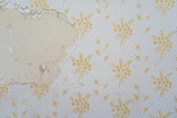 A section of wall features peeling wallpaper with a faded floral pattern. The wallpaper has a beige background with yellow floral designs. Part of the wall is exposed, revealing an uneven, discolored surface underneath.