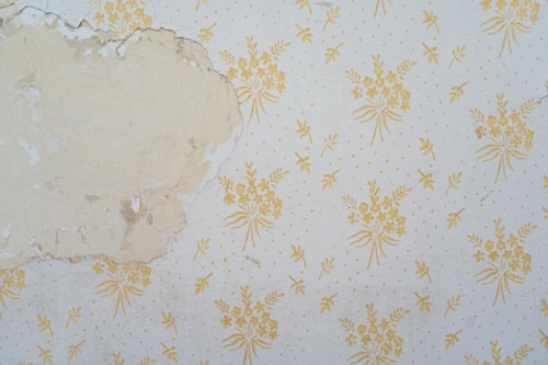 A section of wall features peeling wallpaper with a faded floral pattern. The wallpaper has a beige background with yellow floral designs. Part of the wall is exposed, revealing an uneven, discolored surface underneath.