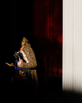 A person wearing a leopard print coat stands partially in shadow while holding a black umbrella, a magazine, and a yellow phone. The background features red lighting against a dark setting with a white column on the right side.