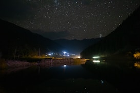 A tranquil night scene with a starry sky overhead, mountains silhouetted against the starlit sky, and a reflective body of water in the foreground. Small patches of light from distant buildings or streetlights create a serene and peaceful ambiance.