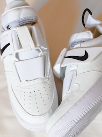 A pair of white sneakers featuring a sleek design with black swoosh logos. They have a modern, strap-based closure system and perforations on the toe area for breathability.