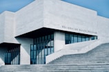 A modern, minimalist building with clean lines and a geometric design. The exterior is dominated by light gray concrete and large glass windows, creating a sleek and contemporary look. The structure features an angular roof and several large steps leading up to the entrance. There is text on the building that appears to be the name of the institution.