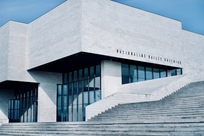 A modern, minimalist building with clean lines and a geometric design. The exterior is dominated by light gray concrete and large glass windows, creating a sleek and contemporary look. The structure features an angular roof and several large steps leading up to the entrance. There is text on the building that appears to be the name of the institution.
