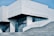 A modern, minimalist building with clean lines and a geometric design. The exterior is dominated by light gray concrete and large glass windows, creating a sleek and contemporary look. The structure features an angular roof and several large steps leading up to the entrance. There is text on the building that appears to be the name of the institution.