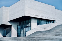 A modern, minimalist building with clean lines and a geometric design. The exterior is dominated by light gray concrete and large glass windows, creating a sleek and contemporary look. The structure features an angular roof and several large steps leading up to the entrance. There is text on the building that appears to be the name of the institution.