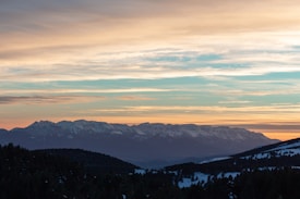 A mountain range is silhouetted against a vibrant sunset, with warm hues of orange, pink, and blue sprawling across the sky. Snow-capped peaks and dark forested hills are visible in the foreground.