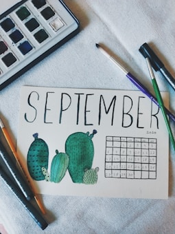 A hand-drawn calendar page for September 2020 featuring green cacti illustrations. Surrounding the page are art supplies including watercolor paints, paintbrushes, and pens.