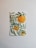 A watercolor painting of orange fruits hanging from green leafy branches. The artwork is mounted on a wall with a light-colored background, highlighting the vibrant colors of the leaves and fruits.