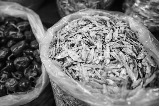 A black and white photograph depicting bags filled with dried fish and another bag containing dark, round objects that resemble dried chili peppers. The bags appear to be made of plastic, arranged closely together, likely at a market setting.