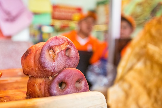 Two pig snouts are displayed prominently in the foreground on a wooden surface, likely at a market or food stall. In the background, there are people wearing orange uniforms, with a blurred and colorful arrangement of objects or decorations.