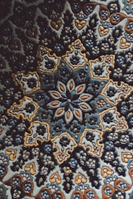 An intricate ceramic tile featuring a symmetrical floral and geometric pattern. The design incorporates repeated motifs of flowers and leaves, predominantly in shades of blue, brown, and white. The craftsmanship highlights a traditional style with complex and detailed artistry.