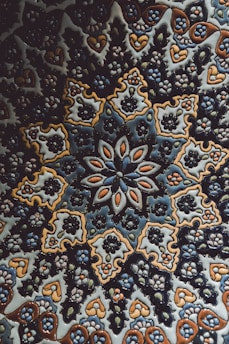 An intricate ceramic tile featuring a symmetrical floral and geometric pattern. The design incorporates repeated motifs of flowers and leaves, predominantly in shades of blue, brown, and white. The craftsmanship highlights a traditional style with complex and detailed artistry.