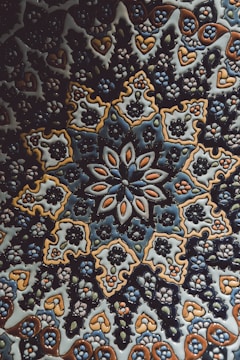 An intricate ceramic tile featuring a symmetrical floral and geometric pattern. The design incorporates repeated motifs of flowers and leaves, predominantly in shades of blue, brown, and white. The craftsmanship highlights a traditional style with complex and detailed artistry.