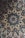 An intricate ceramic tile featuring a symmetrical floral and geometric pattern. The design incorporates repeated motifs of flowers and leaves, predominantly in shades of blue, brown, and white. The craftsmanship highlights a traditional style with complex and detailed artistry.